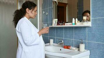 Middle aged multi ethnic pregnant woman in late pregnancy time, standing by white ceramic sink in the home bathroom, drinking water in the morning, dressed in white waffle bathrobe. Healthy lifestyle video