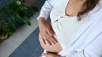 Overhead view of a pregnant woman rubbing moisturizing smoothing body lotion on her belly, to avoid stretch marks in pregnancy, standing at home bathroom interior. Beauty concept. Body and skin care video