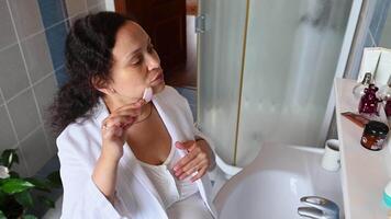 High angle view. Pregnant woman in nightgown and bathrobe using jade stone roller massager, doing lymphatic drainage massage to reduce appearance of first wrinkles and puffiness on her face. Pregnancy video