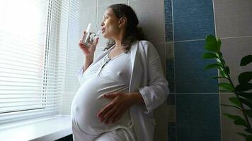 Low angle view of pregnant woman stroking her big belly, drinking water standing by window in the bathroom. Pregnancy. Childbearing. Maternity. Healthy lifestyle. Expecting baby. Morning daily routine video