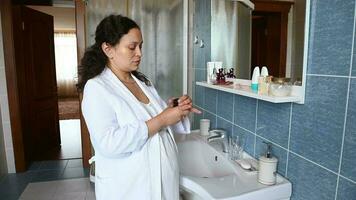 Middle-aged multi ethnic pregnant woman in nightgown and white waffle bathrobe, applying moisturizer on her face, standing in front of a mirror in the home bathroom. Pregnancy time skin care concept video