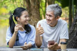 asian teenage girl snap her fingers while listen music together with grandfather in the park on summertime, grandfather doing thumb up for a good song,concept of family lifestyle,the love of a family photo