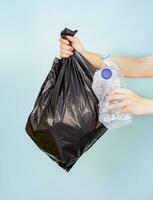 one hand holding a plastic bag filled with plastic bottles, the other holding a plastic bottle waste.isolated on blue background, concept of plastic waste,  waste sorting and waste management photo