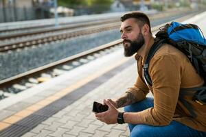 adulto hombre es sentado a ferrocarril estación y esperando para llegada de tren. foto