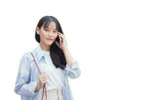 Confident young Asian woman who wears blue white shirt and bag smiles happily while she holds talks to customer on phone with smartphone as she commute to work while isolated on white background. photo