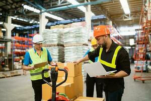 Manager man using laptop check stock and control work with co-worker point at warehouse. Worker wearing high visibility clothing and hard hat, helmet to checking count up goods or boxes for delivery. photo