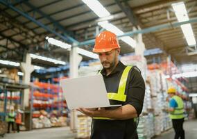 Professional manager man employee using laptop check stock working at warehouse. Worker wearing high visibility clothing and a hard hat, helmet and checking and count up goods or boxes for delivery. photo