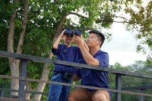 un padre enseña su hijo cómo a utilizar prismáticos a Mira a naturaleza, aves, y fauna silvestre en nacional parques mientras tomando el familia en un cámping día festivo. foto