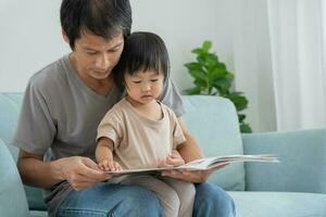 contento asiático padre relajarse y leer libro con bebé hora juntos a hogar. padre sentar en sofá con hija y leyendo un historia. aprender desarrollo, cuidado de niños, reír, educación, contar historias, práctica. foto