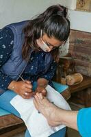 pedicuro haciendo un pedicure a un mayor mujer a hogar durante codicioso 19 pandemia vistiendo cara máscara foto