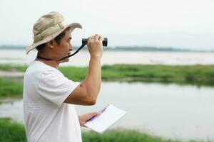 Asian man ecologist is surveying nature at the lake, holds paper notebook and binoculars. Concept, nature exploration. Ecology study.  Pastime activity, lifestyle. Man explore environment photo