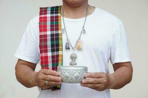 Man wears necklace with Buddha amulets, Thai loincloth on shoulder, holds silver bowl. Concept, faith and belief of Buddhist for holy to protect from dangers, bring good luck, do religious ritual. photo