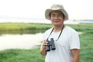 Handsome Asian man ecologist is surveying nature at the lake, holds binoculars.Concept, nature exploration. Ecology study.  Pastime activity, lifestyle. Man explore environment photo