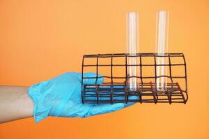 Close up hand wears blue glove, holds test tubes on rack for tube holder, isolated on orange background. Concept, sciecne tool for experiment. Education. Science subject. Learning by doing. photo