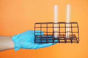 Close up hand wears blue glove, holds test tubes on rack for tube holder, isolated on orange background. Concept, sciecne tool for experiment. Education. Science subject. Learning by doing. photo