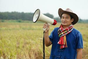 Asian man farmer wears hat, blue shirt, holds megaphone at agriculture land.  Concept, Thai farmer. Agricuture occupation. Protest, announcement. Farmers need help. photo