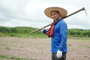 Asian man farmer is at agriculture land, wears hat, blue shirt, holds a hoe on shoulder.Concept organic farming. No chemical. Using traditional manual tool in stead of use herbicide. Zero pollution. photo