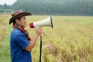 Asian man farmer wears hat, blue shirt, holds megaphone at agriculture land. Concept, Thai farmer. Agricuture occupation. Protest, announcement. Farmers need help. photo