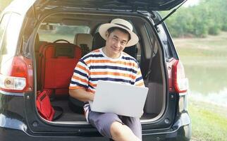 Asian man traveler sits at the trunk of car, working on laptop computer. Concept, outdoor working, freelancer, find inspiration from travel, work and travel. Give time for yourself. photo