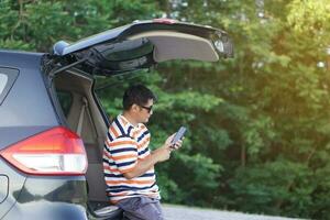 Happy Asian man traveler sits at the trunk of car, using smartphone. Concept, relax time. Vacation, holiday or weekend trip by private vehicle. Transportation. Road trip.Give time for yourself. photo