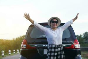 Happy Asian woman traveller wears hat, white shirt and sunglasses, raise hands up to sky at the back of car, feels free. Concept, relax time. Vacation  trip by private vehicle. Give time for yourself. photo