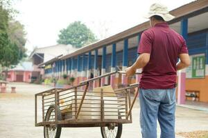 asiático hombre portero usos antiguo carro a llevar cajas a escuela. concepto, ocupación, tailandés portero trabajo y Servicio a escuela, general limpieza de un edificio ,acuerdo colegio mantenido y en bueno condición. foto