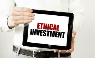 Text ETHICAL INVESTMENT on tablet display in businessman hands on the white background. Business concept photo