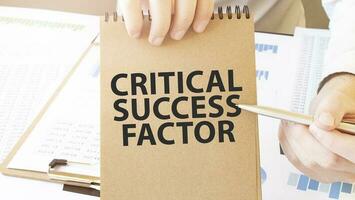 Text CRITICAL SUCCESS FACTOR on brown paper notepad in businessman hands on the table with diagram. Business concept photo