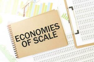 Business up graph on a sheet of craft colour Notepad with ECONOMIES OF SCALE sign. Notepad on desk with financial documentation photo