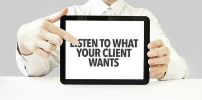 Text LISTEN TO WHAT YOUR CLIENT WANTS on tablet display in businessman hands on the white background. Business concept photo