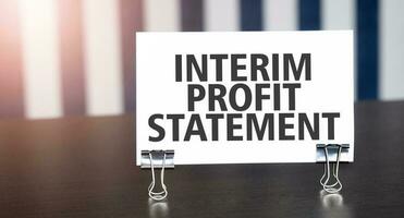 INTERIM PROFIT STATEMENT sign on paper on dark desk in sunlight. Blue and white background photo