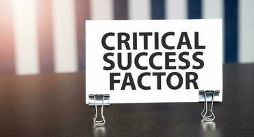 CRITICAL SUCCESS FACTOR sign on paper on dark desk in sunlight. Blue and white background photo