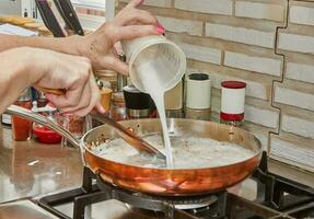 Chef pours milk into chopped onions in copper saucepan over gas fire photo