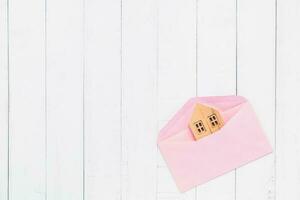 Flat lay of pink envelope with wooden house model on white table photo