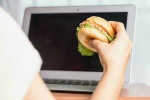 mano participación un delicioso hamburguesa y utilizando ordenador portátil computadora foto