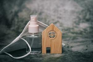 Wooden house model with the bottle of alcohol gel and hygiene face mask on dark background photo