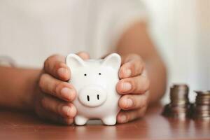Hand holding a piggy bank with blurred coins and money on the wooden table photo