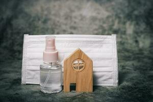 Wooden house model with the bottle of alcohol gel and hygiene face mask on dark background photo