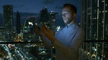 un hombre es utilizando su tableta a noche dentro moderno ciudad Departamento con horizonte ver video