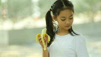 pequeno ásia menina comendo banana dentro a parque. seletivo foco. video