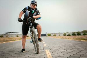 sonriente ciclista en su bicicleta mirando a el cámara en el la carretera. gordito masculino ciclista en ropa de deporte montando un bicicleta al aire libre foto