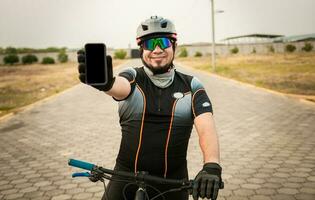 Cyclist in sportswear showing the phone screen. Male cyclist on the bike showing cell phone screen outdoors. Smiling cyclist showing an advertisement on cell phone photo