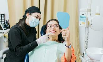 satisfecho hembra cliente en dental clínica mirando a espejo, dentista con paciente sonriente a mano espejo en oficina, hembra paciente comprobación dientes después curación dientes en dental clínica, foto