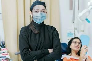 Dental doctor standing in clinic wearing mask, portrait of dentist crossing arms with patient in background, Modern Dental clinic. Dental procedures. portrait of a dentist woman with crossed arms photo