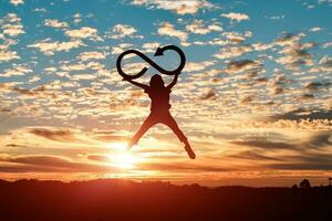 Silhouette of woman jumping and holds an infinity symbol at sunset background. photo