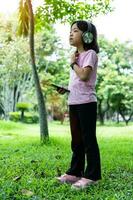 Portrait of child girl with smartphone and listens to music with modern headphones wireless in park outdoors. photo
