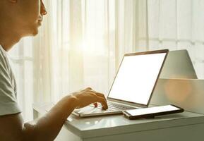 Man's hands using laptop computer with blank screen and smartphone on white table at home. Online shopping, home work, freelance, online learning concept. photo