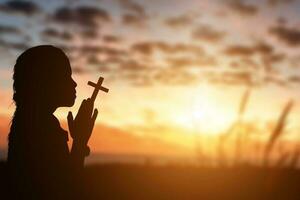 silueta de pequeño niña participación cristiano cruzar en manos a Orando para bendición desde Dios a puesta de sol antecedentes. foto