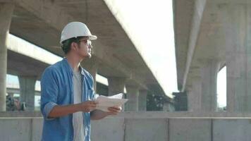 asiatisch jung Fachmann Ingenieur Arbeiter im schützend Helm und Blaupausen Papier auf Hand inspizieren Konstruktion ein Straße Schnellstraße beim konstruieren video