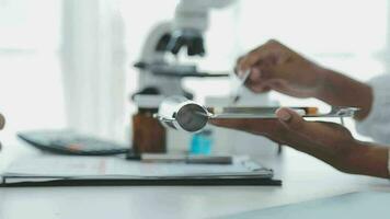 Medical worker in lab coat and sterile mask, doing a microscope analysis while her colleague are working behind video